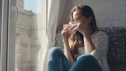 Close-up portrait of charming young woman with frizzy hair sitting in the room enjoying a cup of tea and looking through the window at the city