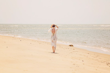 Fototapeta na wymiar Woman in white dress and mala necklace walking away on the sunny beach by the sea