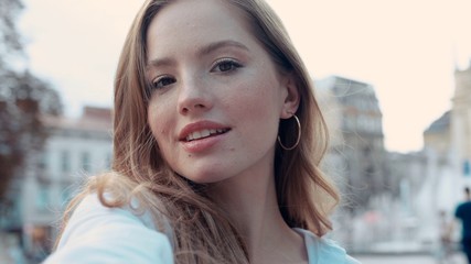 Lifestyle selfie portrait of young cheerful woman with curly golden hair posing and taking selfie on the street smiling and having fun