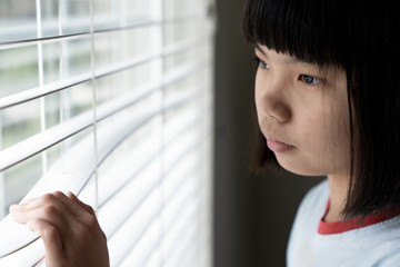 Sad little asian girl quarantee at home, standing near the window