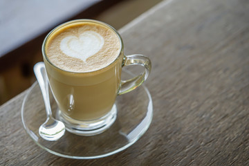 Hot beverage, hot coffee or cappuccino with heart-shaped latte art on top contain in clear cup on clear plate on wooden table with cafe environment