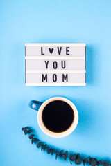 Mom love you written on a decorative panel next to a cup of coffee and eucalyptus on a blue background.