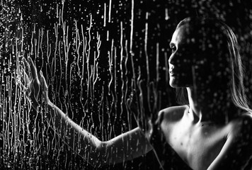 Fototapeta na wymiar A beautiful slim nude girl sensually touches with her hands a window glass, on which streams and drops of rain water flow down on a black background. Artistic monochrome design.