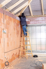 Electrician standing on ladder installing cables in the channel / groove along with electrical boxes in unfinished house built of clay block bricks. Worker pulling cables in house under construction.