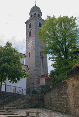 Fototapeta na wymiar Church yard with green trees and stone benches, tall bell tower with cross at the top and clock. Sunny summer day in Lugano, Switzerland.