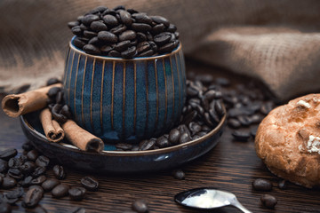 cup full of coffee beans with cinnamon sticks and bun on brown table