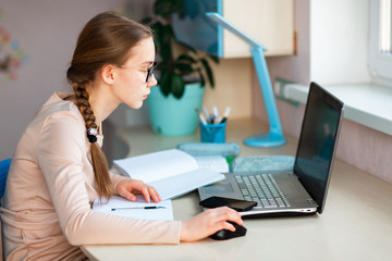 Beautiful young school girl working at home in her room with a laptop and class notes studying in a virtual class. Distance education and learning concept during quarantine