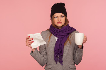 Portrait of irritated flu-sick woman in warm hat and scarf holding tissue and cup of hot beverage, looking with annoyed expression, treating influenza. indoor studio shot isolated on pink background