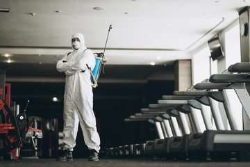 Cleaning and Disinfection in crowded places amid the coronavirus epidemic Gym cleaning and disinfection Infection prevention and control of epidemic. Protective suit and mask and spray bag - obrazy, fototapety, plakaty