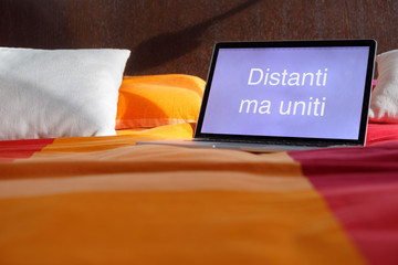 Computer portatile sopra un letto matrimoniale con su scritto: Distanti ma uniti