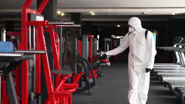 Hazmat worker disinfects gym fitness equipment from coronavirus covid-19 hazard with antibacterial sanitizer sprayer on quarantine. Man in protective suit cleans training apparatus at workout area.