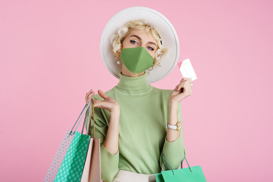 Spring online shopping during quarantine conception: fashionable woman wearing protective mask posing with colorful paper bags and plastic bank card. Pink background. Copy, empty space for text