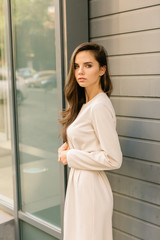 Beautiful brunette young woman wearing white trendy dress and walking on the green street. Spring - summer fashion trend, smiling, happy. 