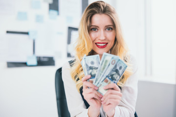 young blond sexy businesswoman manager celebrating successful deal in the end of working day, holding cash like card deck in both hands, welfare concept