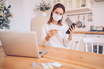 European sick woman in medical mask holding smartphone and thermometer in hands, working on a laptop, kitchen home quarantine isolation Covid-19 pandemic Corona virus. Distance online work, stay home