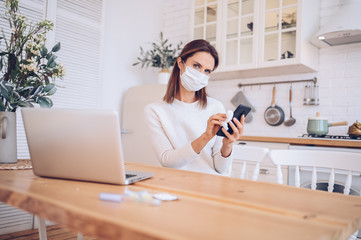 Sick woman in kitchen with laptop cleaning mobile phone by hand sanitizer, using cotton alcohol wool during home quarantine isolation Covid-19 pandemic Corona virus. Distance work from home concept. 