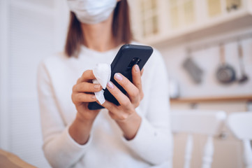 Unrecognizable woman in face protection mask cleaning mobile phone to eliminate germs Covid-19 by hand sanitizer, using cotton wool with alcohol to wipe to avoid contaminating with Corona virus. 