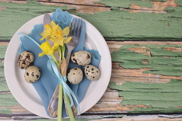 Pretty Easter holiday table setting with cutlery on barn boards. Narcissus, quail eggs, eating utensils with bow on turquoise napkin and white plate. Old green wooden table,top view with copy space.