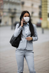 Young and elegant business woman on empty city street using smart phone and wearing protective mask to protect herself from dangerous flu or virus. Corona virus or Covid-19 concept.