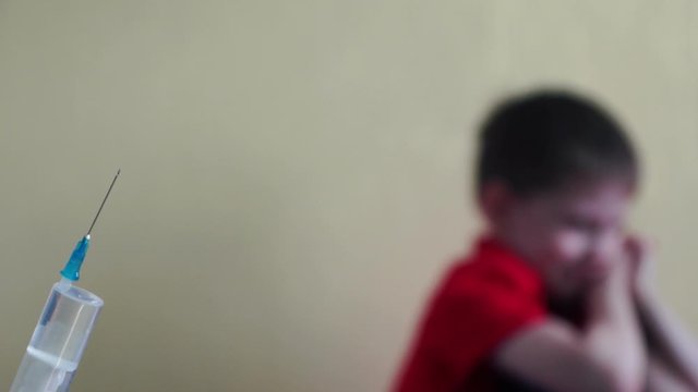 Concept on the topic of vaccination of children. In the foreground a hand holds a syringe, a vaccine. In the background is out of focus a sad boy of 5-6 years old who does not want to give an injectio