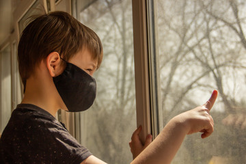 A guy in a protective mask sits at home on isolation and looks out the window while watching the street.