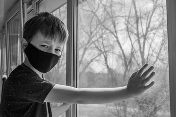 A guy in a protective mask sits at home on isolation and looks out the window while watching the street.