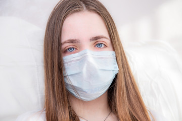 a young girl with brown hair sits at home in quarantine, a young girl sits in bed in a mask from the spread of the virus, infection and looks at the camera. quarantine. stay at home