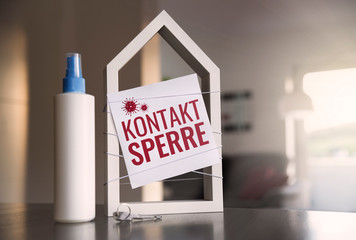 Stilisiertes, weißes Haus mit Label "Kontaktsperre" vor häuslichem Hintergrund mit Desinfektionsspray und Schlüssel