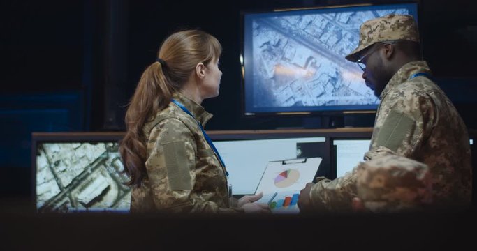 African American man general in camouflage uniform standing in monitoring room and watching troops dislocation on screens. Caucasian woman coming and showing some documents with daa and info.