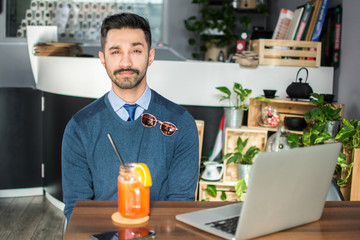 Fototapeta na wymiar Portrait of pretty South Asian young guy with laptop and drink in a cafe