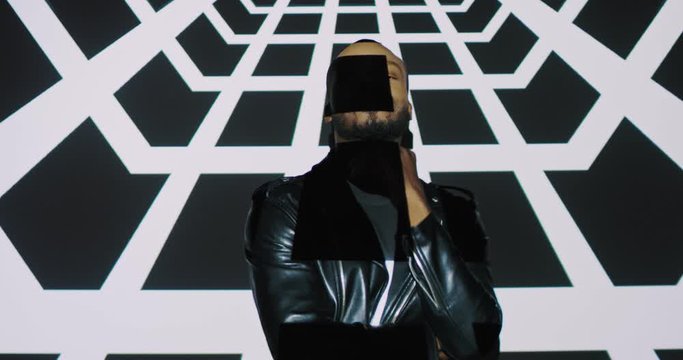 Caucasian attractive young cheerful man in black leather jacket posing to camera and dancing on white wall background with graphic squares projections. Projected figures running on male model.