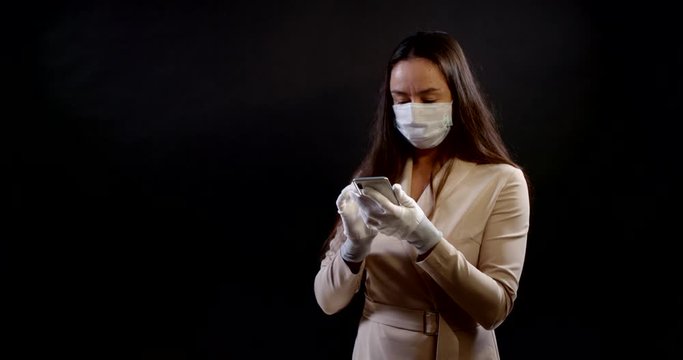 portrait of a woman on a black background. she has put a disposable mask on face and protective gloves on hands, stands three-quarters, holds a smartphone and presses the screen with effort