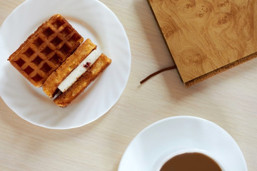 Top view: two pieces of fresh waffles with cream on a plate with morning strong coffee and a notebook on the table