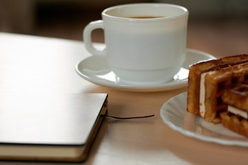 Side view: two pieces of fresh waffles with cream on a plate with morning strong coffee and a closed notebook on the table