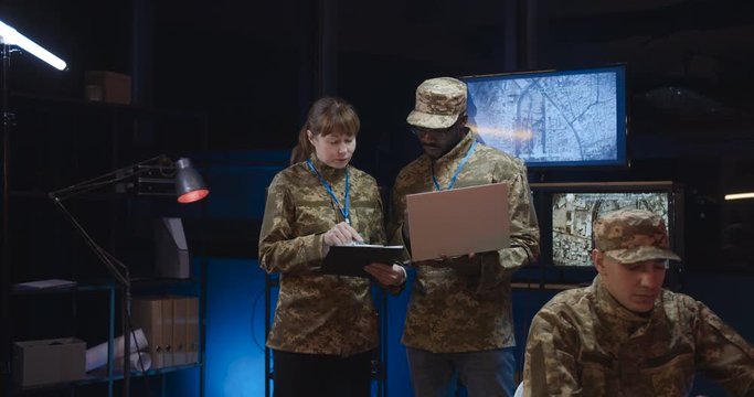 Multiethnic male and female army officials standing in monitoring room and discussing war operation strategy. Caucasian woman with tablet device and African American man with laptop computer.