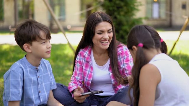 Mom and children sit on the grass and play games on smartphones. Mom's having fun winning her daughter's game and stroking her hair to chear her up. Family concept