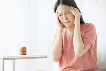 A woman is in a headache sitting on the bed. There is a medicine bottle and a glass of water behind.