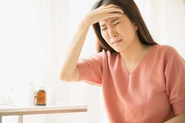 A woman is in a headache sitting on the bed. There is a medicine bottle and a glass of water behind.