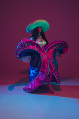 Fototapeta na wymiar Fabulous Cinco de Mayo female dancer on purple studio background in neon light. Beautiful female model in traditional costume and sombrero dancing. Celebration, holiday, beauty and fashion concept.