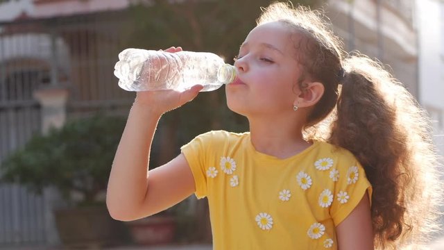 Cute girl quenches thirst, drinks from a plastic bottle on the street near the house. Slow motion little girl eagerly drinks drinking water. Close-up.