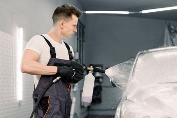 Car wash with flowing water and foam. Side view of young car wash service male employee holding high pressure spray with cleaning foam. Carwash. Washing machine at the station