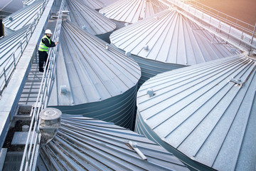 Factory worker in protective clothes walking on grain elevator and observing silos rooftops. Industrial food and grain storage. © littlewolf1989