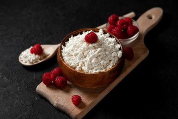 fresh homemade cottage cheese in a wooden plate with raspberries in a rustic bowl against a dark background, healthy food on a black table
