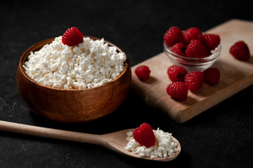 homemade cottage cheese in a wooden plate with raspberries in a rustic bowl against a dark background, healthy food on a black table