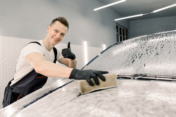 Smiling handsome Caucasian man washing a soapy car hood with a beige sponge, showing ok sign with...
