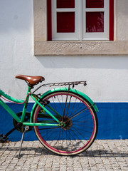 Rueda trasera de una bicicleta retro de color verde y sillín marrón delante de una fachada de una casa blanca y azul