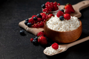 homemade fresh cottage cheese with strawberries, raspberries, blueberries, currants in a wooden plate against a dark background, healthy milk food with wild berries on a black table