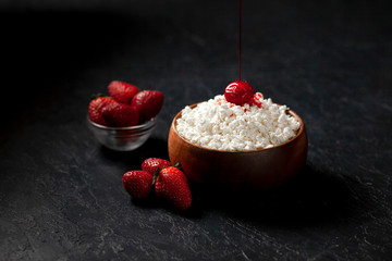 homemade cottage cheese with strawberries and jam in a wooden plate against a dark background, adding red cream to a milk breakfast with berries on a black table