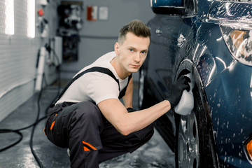Manual car wash in car wash shop service. Handsome young male employee worker cleaning the wheel of...