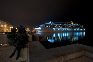 The statues of the seamstresses intent on sewing the Italian flag on the Molo Audace in Trieste, in the background a cruise ship illuminated by the evening lights.